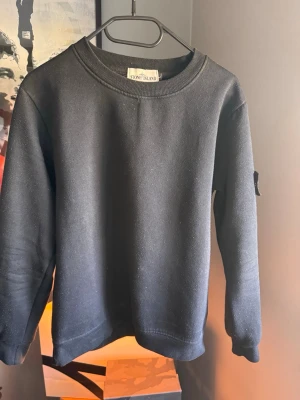 Svart sweatshirt från Stone Island - Svart sweatshirt från Stone Island med klassisk rund halsringning och ribbade muddar. Tröjan har den ikoniska Stone Island-loggan på vänster ärm. Perfekt för en clean och stilren look. Materialet känns mjukt och bekvämt.