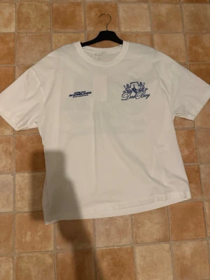 Vit t-shirt med tryck - Vit t-shirt med blått tryck fram och stort motiv med text och fyr på ryggen. Klassisk passform och rund hals. Tillverkad i mjuk bomull, perfekt för en avslappnad stil.
