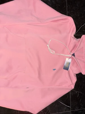 Rosa hoodie från Polo Ralph Lauren - Snygg ljusrosa hoodie från Polo Ralph Lauren med klassisk huva och dragsko. Har en känguruficka framtill och den ikoniska lilla blå loggan broderad på bröstet. Mjuk bomullskänsla och perfekt för en chill och trendig look.