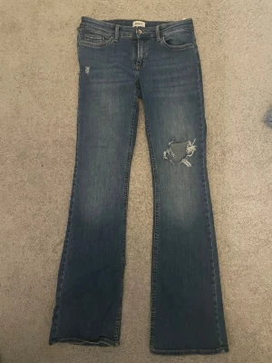 Blå bootcut jeans från ONLY - Snygga blå bootcut jeans från ONLY med slitningar och hål på ena benet. Jeansen har klassisk femficksdesign, normalhög midja och är tillverkade i denim. Perfekta för en avslappnad och trendig look.