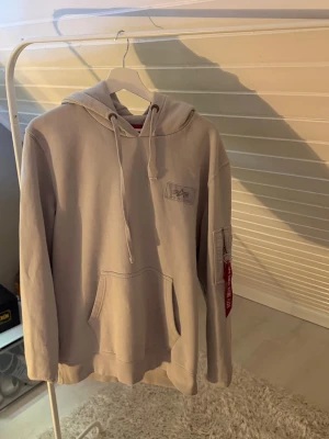 Beige hoodie från Alpha Industries - Snygg beige hoodie från Alpha Industries med klassisk logga på bröstet och praktisk ficka på ärmen. Hoodien har dragsko i huvan och en stor magficka. Perfekt för en chill och avslappnad stil.Hoodien sitter på en kille som väger 80kg och är 190cm lång.