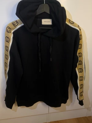 Svart hoodie  - Svart hoodie med huva och dragsnören. Ärmarna har breda beige band med ikoniska GG-loggor och vita detaljer längs sidorna. Klassisk känguruficka framtill. Perfekt för dig som gillar streetstyle och exklusiva märken.