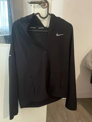 Svart löparjacka från Nike Dri-FIT - Svart löparjacka från Nike med Dri-FIT-material som transporterar bort fukt. Jackan har huva, hel dragkedja framtill och två fickor. Nike-logga i vitt på bröstet och diskret reflexdetalj på ärmen. Perfekt för löpning och träning.