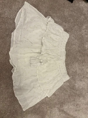 Vita shorts med snörning och volang - Säljer ett par vita shorts med bred resår i midjan, dekorativ snörning med tofsar och volangkant. Shortsen har små diskreta broderade detaljer och är tillverkade i ett lätt och luftigt bomullstyg. Perfekta för varma dagar!