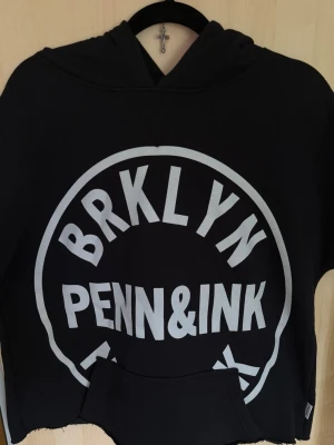 Svart hoodie från Penn&Ink - Cool svart hoodie från Penn&Ink med stor vit BRKLYN PENN&INK-print på framsidan. Klassisk huva och känguruficka framtill. Tillverkad i mjukt material som känns skönt mot huden. Perfekt för en avslappnad streetwear-look.