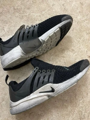 Nike Air Presto svarta sneakers - Nike Air Presto sneakers i svart mesh med grå detaljer och vit sula. Skorna har en sportig design med snörning och plastdetaljer på sidorna. Perfekta för dig som gillar streetwear och vill ha en bekväm och snygg sneaker.