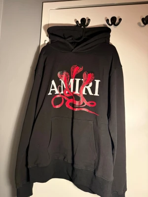 Svart hoodie från Amiri med ormtryck - Svart hoodie från Amiri med stor vit logga och röd ormtryck på bröstet. Hoodien har lång ärm, huva och en stor magficka framtill. Perfekt för dig som gillar streetwear och vill sticka ut med en unik design.