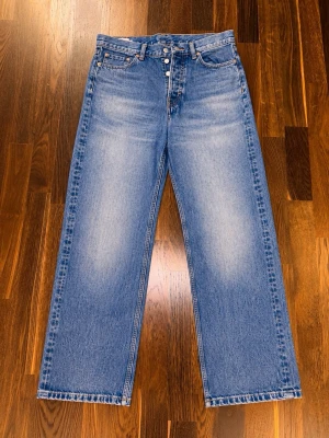 Blå raka jeans med hög midja - Säljer ett par klassiska blå jeans med raka ben och hög midja. Jeansen har fem fickor, knäppning med knappar och snyggt ljus tvättad denim. Perfekta för en avslappnad och trendig look.