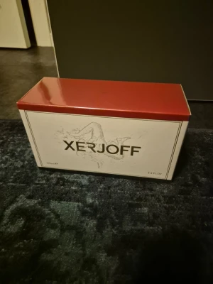 Xerjoff parfym 100ml exklusiv - Lyxig parfym från Xerjoff i en elegant rektangulär kartong med röd ovansida och vita sidor. Förpackningen rymmer 100 ml och har stilren design med Xerjoff-loggan i svart. Perfekt för dig som gillar exklusiva dofter.
