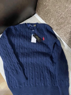 Mörkblå kabelstickad tröja Polo Ralph Lauren - Mörkblå kabelstickad tröja från Polo Ralph Lauren med rund halsringning och klassisk röd broderad logga på bröstet. Tröjan har långa ärmar och ribbade muddar. Perfekt för dig som gillar stilren och tidlös design.