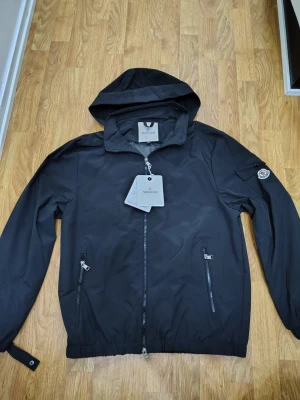 Svart vindjacka från Moncler - Snygg svart vindjacka från Moncler med huva och dragkedja framtill. Jackan har två framfickor med dragkedja, elastiska muddar och en Moncler-logga på ärmen. Tillverkad i lätt, vattenavvisande material som passar perfekt för blåsiga dagar.