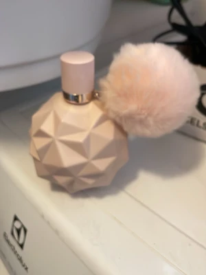 Ariana Grande parfym med fluffig boll - Superfin parfym från Ariana Grande i en ljusrosa, geometrisk flaska med en stor fluffig boll som detalj. Flaskan har en modern design och guldig ring vid halsen. Perfekt för dig som gillar söta och trendiga dofter.