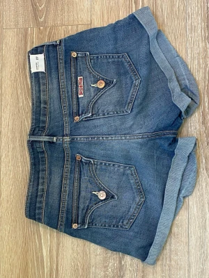 Blå jeansshorts från Elton - Snygga blå jeansshorts från Elton med uppvikta benslut och klassiska fem fickor. Shortsens bakfickor har dekorativa sömmar och en liten brittisk flagga. Perfekta för en avslappnad och trendig look.