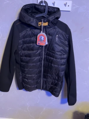 Svart pufferjacka från Parajumpers - Snygg svart pufferjacka från Parajumpers med huva och dragkedja framtill. Jackan har quiltad front, stickade ärmar och axlar samt en bröstficka med dragkedja. Perfekt för kalla sommardagar och riktigt trendig look.