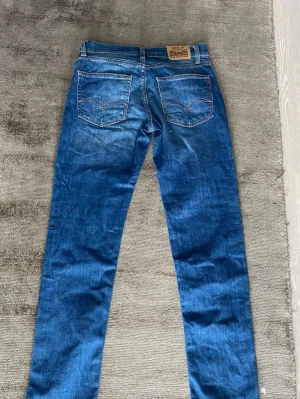Blå jeans från Replay - Säljer ett par klassiska blå jeans från Replay med raka ben och snygga detaljer på bakfickorna. Jeansen har en läderpatch med Replay-logga bak i midjan och är tillverkade i slitstark denim. Perfekta för en avslappnad och trendig look.