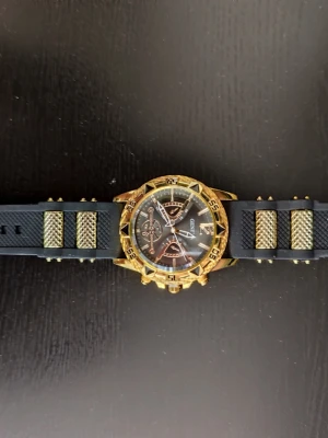 Svart och guld klocka från Geneva - Stilren klocka från Geneva med svart urtavla och detaljer i guld. Klockan har ett sportigt utseende med svart armband och guldiga inslag. Urtavlan har flera dekorativa visare och siffror, och boetten är markerad med guld och svarta detaljer. Ända att klaga på är att minutvisaren är lite lös när man vrider på klockan 