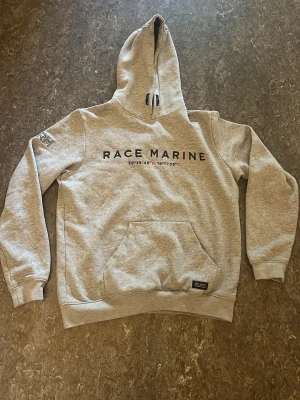 Grå hoodie från Race Marine - Snygg grå hoodie från Race Marine med tryck på bröstet och broderad RM-detalj på ärmen. Klassisk känguruficka framtill och justerbar huva med snörning. Perfekt för en avslappnad och sportig stil.