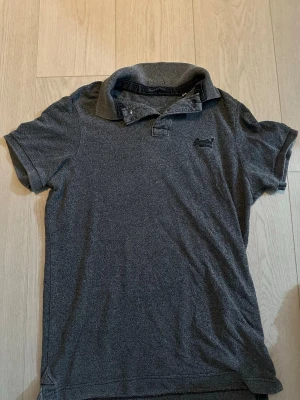 Mörkgrå pikétröja från Superdry - Snygg mörkgrå pikétröja från Superdry med klassisk krage och korta ärmar. Tröjan har broderad logga på bröstet och är tillverkad i ett mjukt och bekvämt material. Perfekt för en avslappnad och stilren look.