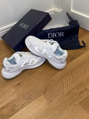 Dior b30 stolek 41 - Säljer ett par snygga vita Dior sneakers med reflekterande detaljer och chunky sula. Skorna har mesh och syntetmaterial i ovandelen, samt coola silverinslag och snörning. Perfekta för dig som vill ha en exklusiv och trendig look.