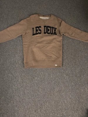 Beige sweatshirt från Les Deux - Snygg beige sweatshirt från Les Deux med svart broderad text på bröstet. Tröjan har rund halsringning, långa ärmar och ribbade muddar. Perfekt för en avslappnad och trendig stil.