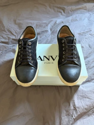 Svarta Lanvin sneakers i läder och canvas - Pris kan ej diskuteras… Snygga svarta sneakers från Lanvin med ovandel i canvas och tåparti i läder. Skorna har vita sulor och svarta skosnören, samt diskreta metalldetaljer vid snörningen. Perfekta för dig som gillar stilrena och exklusiva sneakers.