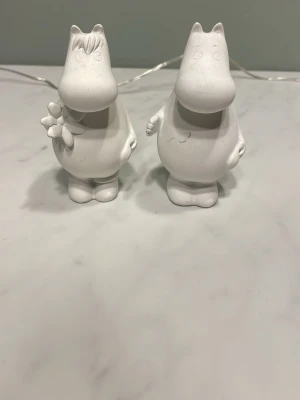 Muminfigurer i vitt keramik - Två vita Muminfigurer i keramik, en med blomma och lugg och en utan detaljer. Figurerna har rund form och står upprätt. Perfekta som dekoration eller samlarobjekt för Mumin-fans.
