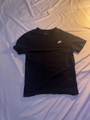 Svart Nike t-shirt med logga - Svart t-shirt från Nike med klassisk vit logga på bröstet. Enkel och stilren design med rund hals och korta ärmar. Perfekt för en sportig och avslappnad look.