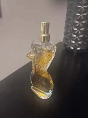 Jean Paul Gaultier Classique parfym - Ikonisk parfymflaska från Jean Paul Gaultier i form av en kvinnlig byst. Flaskan är genomskinlig med guldigt spruthuvud och innehåller en bärnstensfärgad vätska. Perfekt för dig som gillar unika och snygga designer på parfymflaskor.