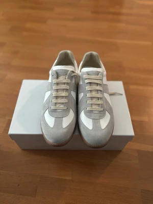 Maison Margiela Gats White - Snygga sneakers i grå mocka och vitt läder med beige snörning och klassisk gummisula. Modellen har en retroinspirerad design som passar perfekt till streetwear och casual outfits. Låg profil och stilrena detaljer gör dem lätta att matcha.