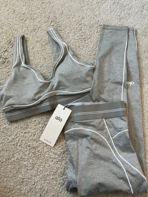 Grått träningsset från Alo Yoga - Snyggt grått träningsset från Alo Yoga med sport-bh och matchande tights. Setet har vita kontrastsömmar och bred resår i midjan och under bysten. Materialet är stretchigt och passar perfekt för gym eller yoga.