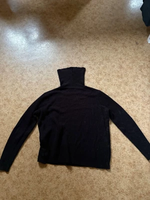 Svart turtleneck i stickat material - En svart stickad turtleneck med lång ärm och hög krage. Tröjan har en klassisk rak passform och är perfekt för lager-på-lager-stil. Materialet är mjukt och ribbstickat för extra komfort.