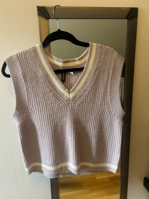 Stickad väst med v-ringning - Säljer en ljus lila stickad väst från H&M med v-ringning och ribbade kanter. Västen har ljusa ränder vid halsringning och nederkant, vilket ger en fräsch och trendig look. Perfekt att styla över en skjorta eller t-shirt. Helt ny utan prislapp 