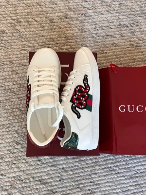 Gucci Ace sneakers med orm - Gucci Ace sneakers i vitt skinn med ikonisk broderad orm på sidan, gröna och röda detaljer samt klassisk snörning. Skorna har metallic-detaljer på hälen i grönt och rött och den typiska Gucci-randen i grönt och rött på sidan. Perfekta för dig som vill sticka ut. Hör av dig innan du köper!