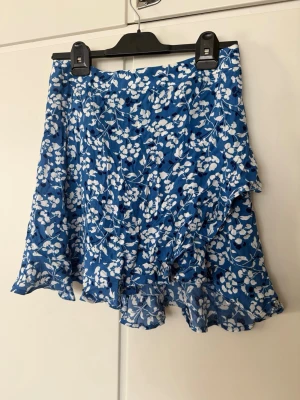 Blå blommig volangkjol - Supersöt blå kortkjol med vitt blommönster och volangdetaljer. Kjolen har omlottkänsla och är tillverkad i ett lätt och luftigt material, perfekt för varma dagar. Passar dig som vill ha en feminin och trendig look.