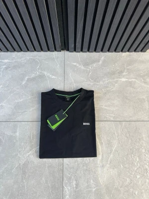 Svart BOSS t-shirt med logga - Snygg svart t-shirt från BOSS med klassisk passform och diskret vit logga på bröstet. T-shirten har rund halsringning och gröna detaljer på insidan av kragen. Perfekt basplagg för en stilren look.