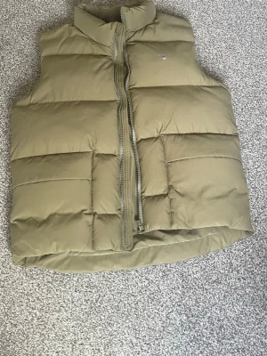 Olivgrön dunväst från GANT - Snygg olivgrön dunväst från GANT med hög krage och dragkedja framtill. Västen har två stora fickor och quiltad design som ger en modern look. Perfekt för lager-på-lager-stil under kyliga dagar. Nypris: 1600. Storlek: 158-164 helt oanvänd, prislapp på. Öppen för diskutering om prisk