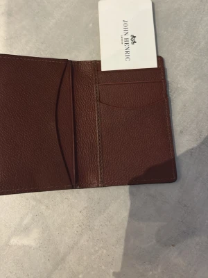 Brun läderplånbok John Henric - Slimmad brun plånbok i äkta läder från John Henric. Plånboken har flera kortfack och stilren design med präglad logotyp på framsidan. Perfekt för dig som vill ha en smidig och snygg accessoar i fickan.