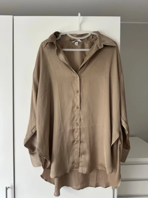 Oversized beige skjorta i satin - Stilren oversized skjorta från H&M i beige satinliknande material. Skjortan har klassisk krage, knäppning framtill och långa ärmar med breda manschetter. Perfekt för en avslappnad men trendig look. Har fläck som man kan se på bild 2