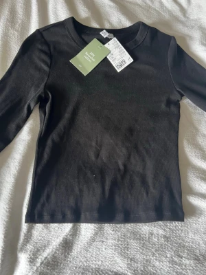 Svart ribbad långärmad topp H&M - Svart ribbad långärmad topp från H&M med rund halsringning. Tillverkad i mjuk bomullsblandning med 25% återvunnen bomull. Enkel och stilren modell som passar till många olika outfits.