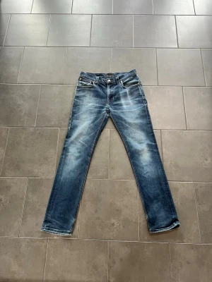 Nudie jeans herr - Snygga blå jeans med raka ben och tydliga slitningar framtill för en avslappnad look. Klassisk femficksmodell med kontrastsömmar och normal midja. Perfekta till sneakers och hoodie.