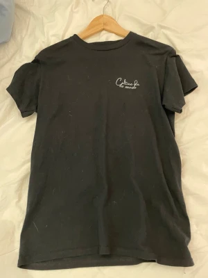Svart t-shirt Culture de la mode - Svart t-shirt från Culture de la mode med tryck på bröstet och en stor grafisk print på ryggen. Klassisk rund hals och korta ärmar. Tillverkad i mjuk bomull för en skön känsla och avslappnad stil.
