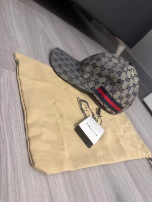 Blå/grå canvas keps - Snygg keps med klassiskt monogram i  marinblått. Kepsen har en röd och marinblå rand på sidan och justerbar rem baktill. Materialet är canvas.