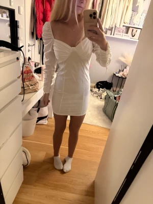 Vit kort klänning med puffärmar - Snygg vit kort klänning med v-ringad front och puffiga långärmar. Klänningen har figurnära passform och markerad byst, perfekt för dig som vill ha en trendig och stilren look. 