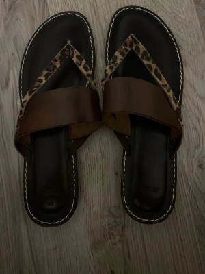 Bruna sandaler med leoparddetaljer - Snygga sandaler med breda bruna remmar och leopardmönstrade detaljer. Brun sula med vit söm runt kanten ger en stilren kontrast. Perfekta för en trendig och avslappnad look.