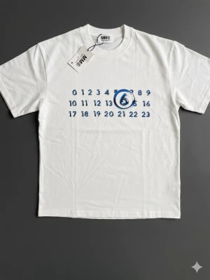 Vit t-shirt från MM6 Maison Margiela - Vit t-shirt från MM6 Maison Margiela med tryck av blå siffror och en stor blå cirkel runt siffran 6 på bröstet. Klassisk rund hals och korta ärmar. Tillverkad i mjuk bomull för en bekväm känsla. Perfekt för dig som gillar minimalistisk och modern design.