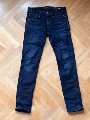 Mörkblå slim jeans från Replay Anbass - Snygga mörkblå slim fit jeans från Replay med klassisk femficksdesign och coola detaljer på bakfickorna. Jeansen har en smal passform och är tillverkade i stretchigt denimtyg för extra komfort. Perfekta för en trendig och avslappnad stil.