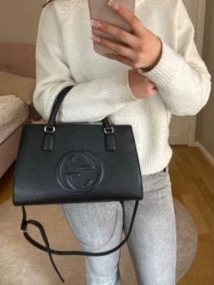 Svart handväska från Gucci ( Gucci Soho bag)  - Snygg svart handväska från Gucci i strukturerat läder med präglad logga framtill. Väskan har både handtag och justerbar axelrem i samma färg, samt guldfärgade metalldetaljer. Perfekt storlek för det viktigaste och ger en lyxig touch till din outfit. Väskan är en vintage väska som bara använts några gånger, den är i mycket bra skick. 