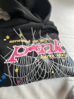 Svart hoodie från Spider, M - Cool svart hoodie från Spider med färgglatt tryck framtill. Texten 'never mind the spider' och 'here's the pink' i gult, rosa och vitt, samt stjärnor och spindelnätsmönster. Perfekt för dig som gillar unika och lekfulla plagg. Huva och mjukt material.