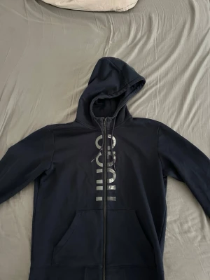 Mörkblå HUGO zip hoodie - Snygg mörkblå hoodie från HUGO med dragkedja framtill och stor huva. Stora fickor på magen och ett vertikalt HUGO-tryck i silver på bröstet. Perfekt för en avslappnad och trendig look. Tillverkad i mjukt bomullsmaterial.