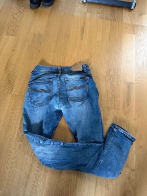 Blå grim Tim jeans från nudie  - Säljer ett par klassiska blå jeans från Nudie Jeans med snygga slitningar och kontrastsömmar. Modellen har raka ben, fem fickor och coola detaljer på bakfickorna. Perfekta för en avslappnad och trendig look.